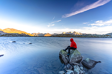 Hiker sitting on rocks on water edge of Laghi Azzurri (Bergsee) at dawn, Spluga Pass, Valle Spluga, Valtellina, Lombardy, Italy, Europe