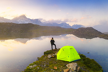 Rear view of man with hat standing outside tent at Obere Schwarziseeli lake at dawn, Furka Pass, Canton Uri, Switzerland, Europe