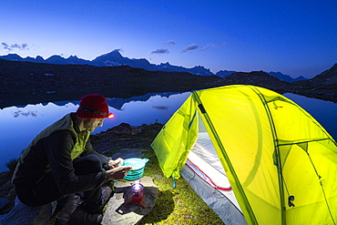 Hiker man cooking outside camping tent at Obere Schwarziseeli lake at dusk, Furka Pass, Canton Uri, Switzerland, Europe