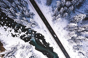 Car traveling on snowy mountain road across frozen river and woods, aerial view, Switzerland, Europe