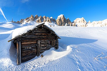 Isolated wooden hut covered with snow with Cir Group peaks in background at sunset, Passo Gardena, Dolomites, South Tyrol, Italy, Europe