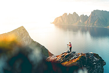 Photographer with tripod on Husfjellet mountain watching sunset on sharp peaks of mount Okshornan (Devil's Teeth), Senja, Norway, Scandinavia, Europe