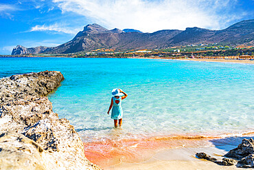 Woman in dress standing in the turquoise clear sea facing the pink sand beach of Falassarna, Crete, Greek Islands, Greece, Europe