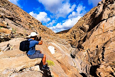 Tourist photographing canyons with smartphone sitting on rocks at Barranco de las Penitas, Fuerteventura, Canary Islands, Spain, Atlantic, Europe