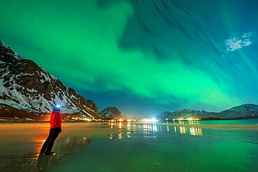 Hiker with head torch watching the Northern Lights standing on Ramberg beach, Nordland county, Lofoten Islands, Norway