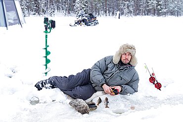 Man lying on ice while fishing from a hole, Lapland, Sweden