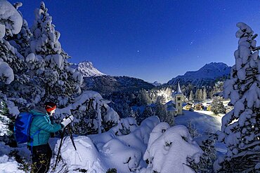 One man photographing the old Chiesa Bianca at night standing in a snowy forest, Maloja, Graubunden, Engadin, Switzerland