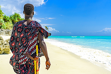 Maasai man with traditional clothing and stick admiring the crystal sea standing on a beach, Zanzibar, Tanzania, East Africa, Africa