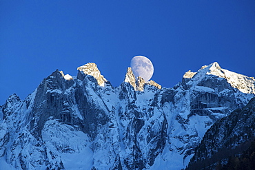 The moon appears behind the snowy mountains illuminating the peaks at sunset, Bondasca Valley, Swiss Alps, Switzerland, Europe