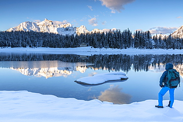 Hiker admires the snowy peaks and woods reflected in Lake Palu at dawn, Malenco Valley, Valtellina, Lombardy, Italy, Europe