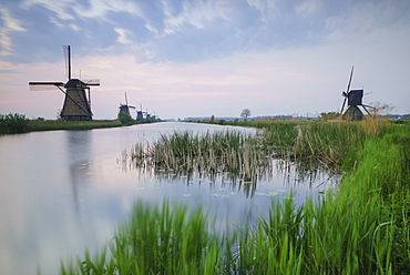 Green grass frames the windmills reflected in the canal at dawn, Kinderdijk, Rotterdam, South Holland, Netherlands, Europe