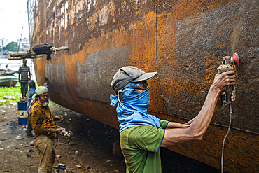 Men repair a boat in the shipwreck yard, focusing on the hull and using tools to remove rust and old paint at a worksite in Bangladesh