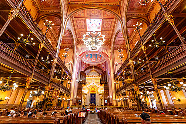 Symmetrical wide-angle interior view of Dohány Street Synagogue showing chandeliers, balconies, and the ark beneath painted ceiling. Horizontal landscape orientation.