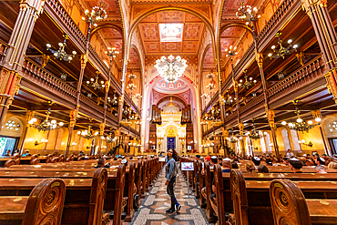 Wide-angle interior view of Dohány Street Synagogue, showing pews, chandeliers, ark, and a visitor standing in the aisle. Horizontal landscape orientation.