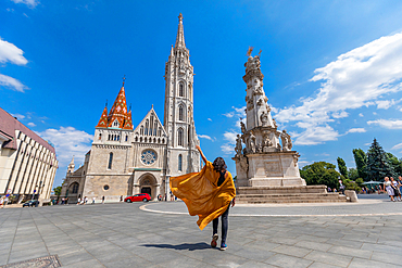 Street-level wide-angle shot of a traveler in yellow coat with flowing fabric approaching Matthias Church and Holy Trinity Column. Horizontal landscape orientation.