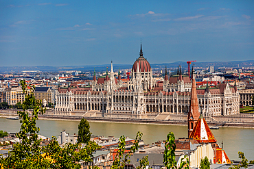 Elevated wide-angle view of the Hungarian Parliament Building on the Danube riverbank, seen from Buda with surrounding rooftops. Horizontal landscape orientation.