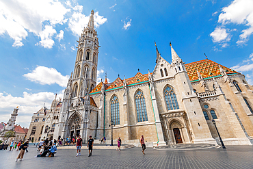 Wide-angle ground-level view of Matthias Church tower and facade, surrounded by visitors in the Castle District square on a sunny day. Horizontal landscape orientation.