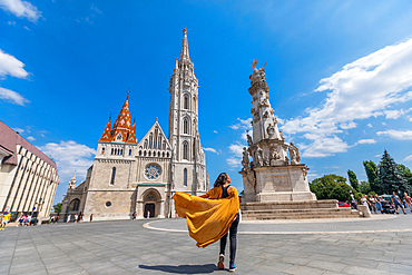 Wide-angle street view of a traveler in a flowing yellow coat walking toward Matthias Church and the Holy Trinity Column in Budapest. Horizontal landscape orientation.