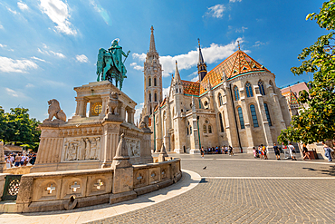 Wide-angle ground view of Matthias Church with its colorful tiled roof and the equestrian statue of St. Stephen in Budapest’s Castle District. Horizontal landscape orientation.