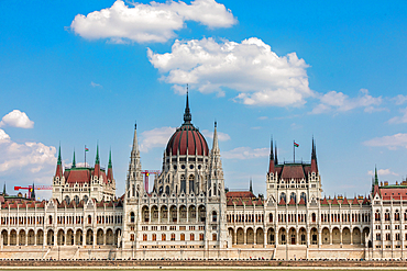 Frontal wide-angle view of the Hungarian Parliament Building on the Danube riverbank, with Gothic Revival architecture under a blue sky. Horizontal landscape orientation.