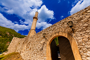 Ground-level upward view of stone fortress walls with arched entrance and tall mosque minaret against a blue sky. Horizontal landscape orientation.
