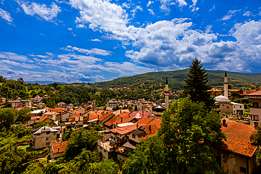 Wide-angle elevated view over the red rooftops and mosques of Travnik surrounded by rolling green hills and mountains. Horizontal landscape orientation.