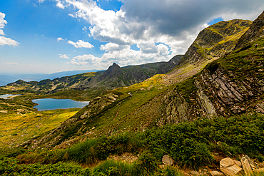 Horizontal, elevated view — Multiple mountain lakes in Rila Mountains, Bulgaria. Rocky slope in foreground, distant peaks under partly cloudy sky.