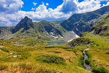 Horizontal, eye-level shot – Meadow and small stream in foreground with peaks and lake in background. Rila Mountains, Bulgaria.
