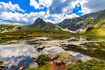 Horizontal, eye-level shot – Lake surface reflecting peaks and clouds, with alpine vegetation in foreground. Rila Mountains, Bulgaria.
