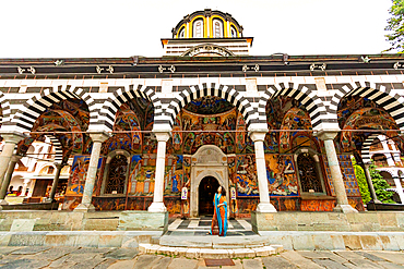 Horizontal, eye-level shot of the main church entrance at Rila Monastery in Rila, Bulgaria, showing striped arches, colorful frescoes, and a person standing in the doorway.