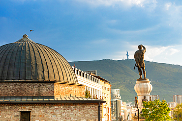Horizontal eye-level shot of a domed stone building and a large statue of a man holding a sword, set against a backdrop of mountains and a cloudy blue sky in Skopje, North Macedonia.