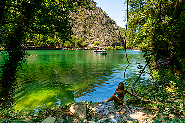 Horizontal eye-level shot framed by overhanging trees, showing a woman sitting at the water’s edge of Matka Canyon with emerald-green waters, a tour boat in the distance, and steep rocky cliffs in the background.
