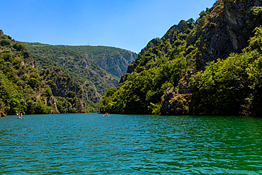 Horizontal eye-level shot of Matka Canyon’s turquoise waters flanked by steep, rocky cliffs covered in dense green vegetation under a clear blue sky.