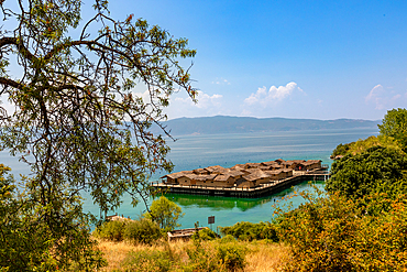 Horizontal shot from an elevated viewpoint, partially framed by tree branches, showing a stilted village of thatched huts over turquoise water, with golden dry grass and shrubs in the foreground and distant mountains across the lake.