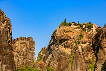 Horizontal ground-level shot capturing a monastery atop a high rock formation, framed by surrounding vertical cliffs under a clear blue sky. The angle emphasizes the height and isolation of the structure without dramatic embellishment.
