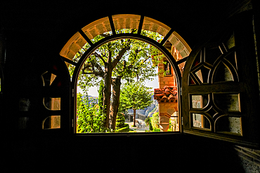 This image captures a scenic view framed by a large arched wooden window with open shutters. Through the window, you can see a sunlit courtyard with a tree, part of the monastery's tiled roof, and distant mountains under a bright sky.
