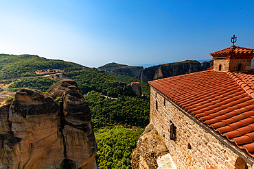 A red-tiled roof of a Meteora monastery extends to the cliff's edge, overlooking vast green valleys and dramatic rock formations under a clear blue sky.