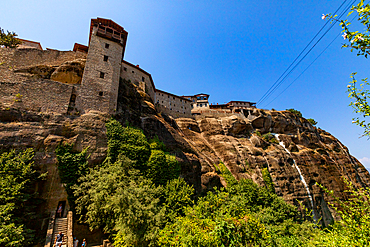 Side view of the Holy Monastery of Varlaam in Meteora, with its stone tower, cliffside structures, and a waterfall cascading down the rock face.