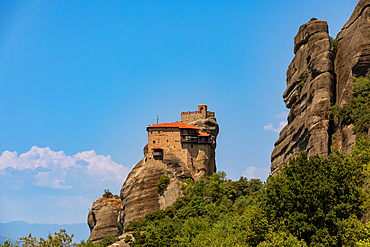 The Holy Monastery of Rousanou, dramatically situated on a narrow rock pillar, with its stone walls and red-tiled roof standing out against the rugged cliffs and bright blue summer sky in Meteora, Thessaly, Greece.
