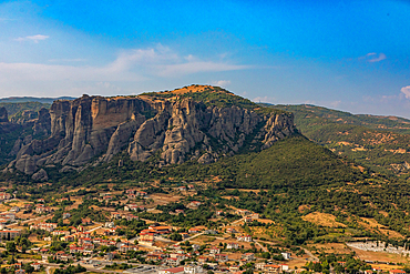 Landscape orientation view of a town below a rocky cliff, with a grassy hill and scattered trees rising in the background, Thessaly, Greece.