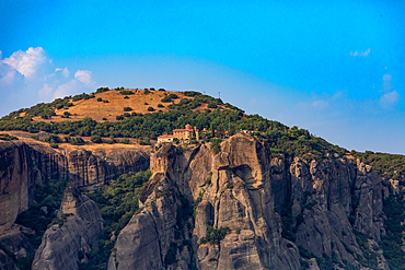 Landscape orientation view of a Meteora monastery perched atop rocky cliffs, with a hilltop covered in grass and trees in the background, Thessaly, Greece.