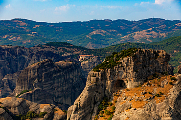 Aerial view in landscape orientation of a Meteora monastery perched on a towering rock pillar, with rugged cliffs and forested hills in the background, Thessaly, Greece.