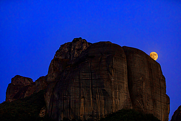 Landscape orientation view of the full moon rising behind towering rock formations at Meteora, a UNESCO World Heritage site in Thessaly, Greece.