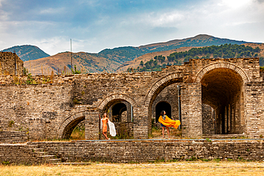 Landscape orientation view of two women in colorful dresses posing among the stone archways of Gjirokastër Castle, with mountain scenery in the background, Gjirokastër, Albania.