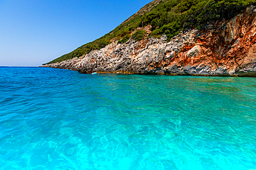 Clear turquoise waters of the Ionian Sea meeting rocky, vegetation-covered coastline near Dhermi, Vlorë County, Albania, on a bright summer day.
