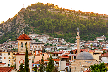 A mosque and church rise above the rooftops of Berat, Albania, with the hillside Kala Castle and rows of Ottoman-style houses in the background at sunset.