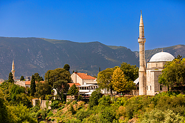 The Koski Mehmed Pasha Mosque rises above the rooftops of Mostar’s Old Town, framed by lush greenery and the rugged Dinaric Alps under a clear blue sky.
