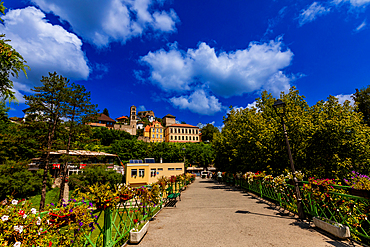 A flower-adorned pedestrian bridge in Jajce, Federation of Bosnia and Herzegovina, Bosnia and Herzegovina, leading to the old town with views of the Saint Luke bell tower and historic buildings under a bright summer sky.