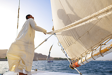 An Egyptian man stands on the bow of a traditional Felucca sailboat with wooden masts and cotton sails on the River Nile, Aswan, Egypt, North Africa, Africa