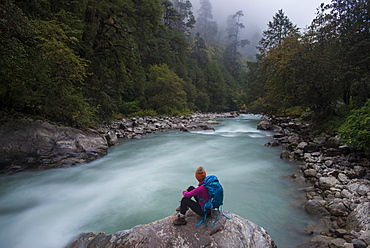 A woman takes a break from the trail and sits beside the Langtang Khola near the little village of Riverside on a misty evening, Nepal, Himalayas, Asia